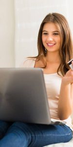 Young smiling female making an online payment using a laptop which is placed on her lap holding a credit card in hand